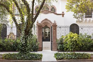 Ornate doorway of a white building with decorative stonework, wrought iron gate, and large arched windows, surrounded by lush green trees and shrubs along a sidewalk.