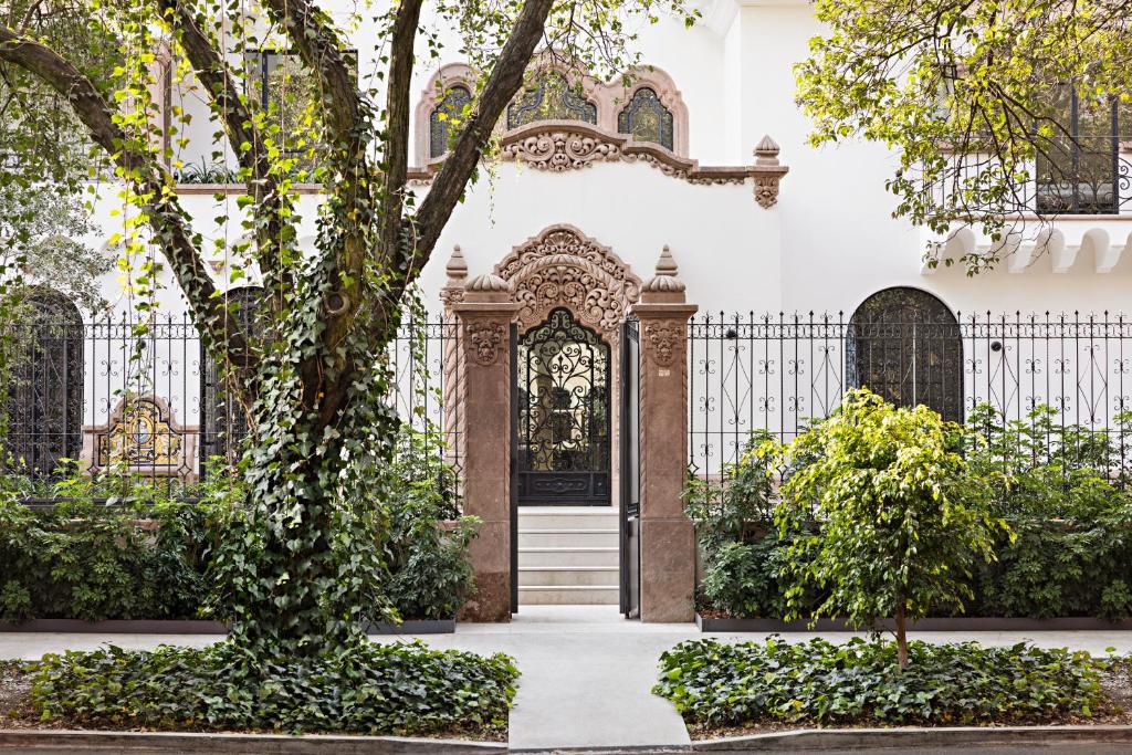 Ornate doorway of a white building with decorative stonework, wrought iron gate, and large arched windows, surrounded by lush green trees and shrubs along a sidewalk.