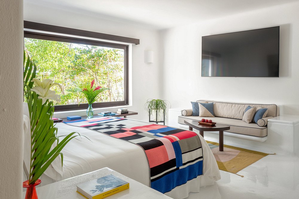 A bright, modern bedroom with a large window showing greenery, a bed covered with a colorful geometric blanket, a sofa, a coffee table, a large wall-mounted TV, and fresh flowers in vases.