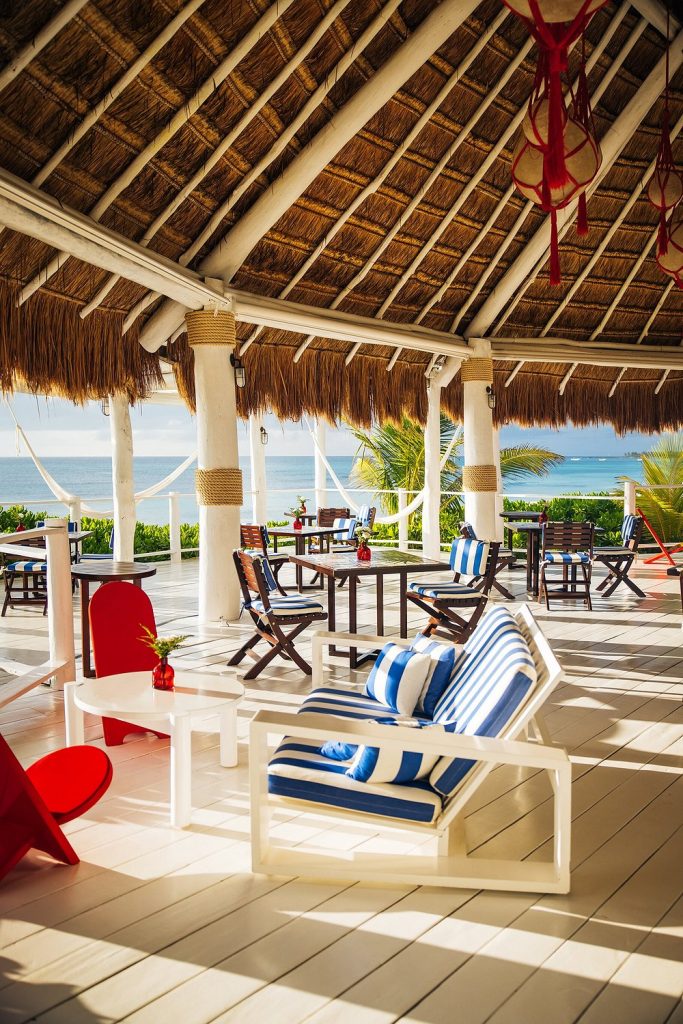 Open-air beachside lounge with a thatched roof, featuring white furniture with blue-striped cushions, wooden chairs, red accents, and a view of the ocean and palm trees in the background.