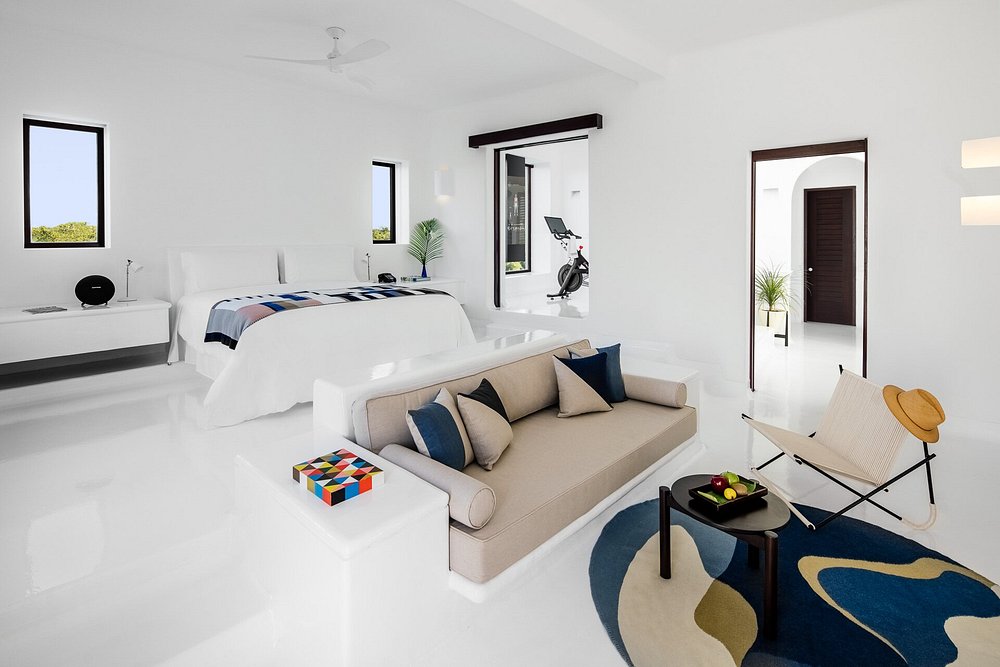 Spacious, modern bedroom with white walls and floor, featuring a bed, beige sofa, chair with hat, colorful rug, and exercise bike near a window. Two doorways lead to other rooms with plants and natural light.