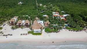 Aerial view of a beachfront resort with thatched-roof buildings and villas surrounded by lush greenery, set along a white sand beach with lounge chairs and umbrellas facing the ocean.