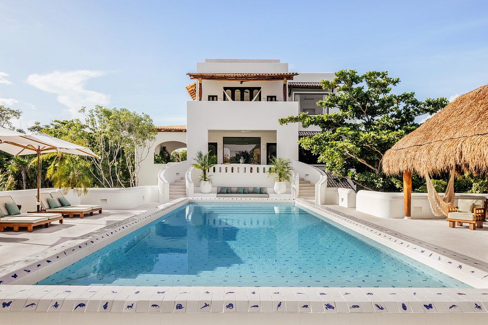 A luxurious white villa with a large outdoor swimming pool, surrounded by lounge chairs, umbrellas, lush greenery, and a thatched-roof seating area, under a clear blue sky.