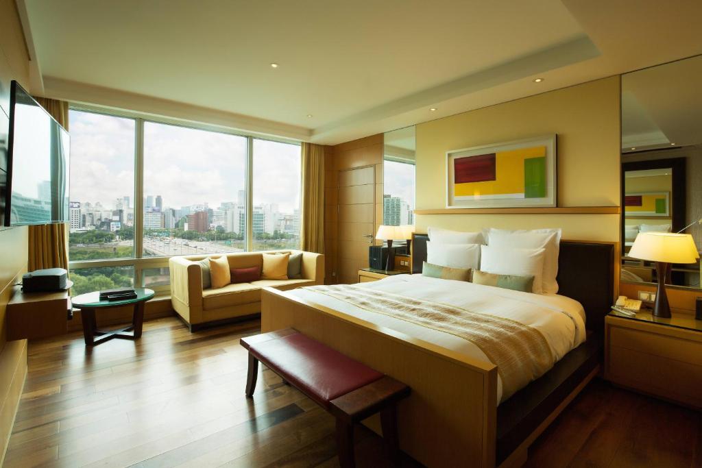 Modern hotel room with large windows showing a city view, a neatly made bed, a beige sofa, wooden floor, a wall-mounted TV, a desk, and warm lighting, creating a cozy and inviting atmosphere.