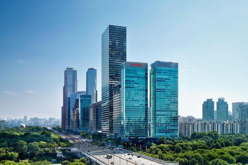 A cityscape with tall modern skyscrapers, including several glass office buildings, overlooking a busy road and lush green trees under a clear blue sky.