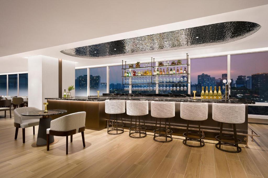 A modern, stylish bar with a marble countertop, high-backed chairs, and backlit shelves displaying bottles and glassware, set against large windows overlooking a city skyline at sunset.