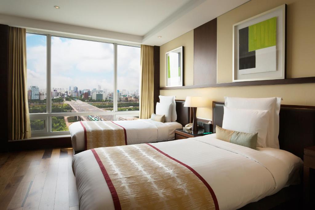 A modern hotel room with two neatly made single beds, large window with a city view, hardwood floor, bedside lamp, and contemporary artwork on the walls.