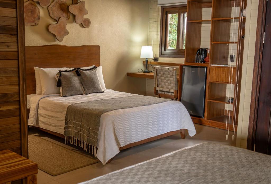 A cozy bedroom with a neatly made bed, gray throw blanket, and decorative pillows. There is a wooden headboard, wall art, a lamp on a small desk, a mini fridge, and shelves near a window with natural light.