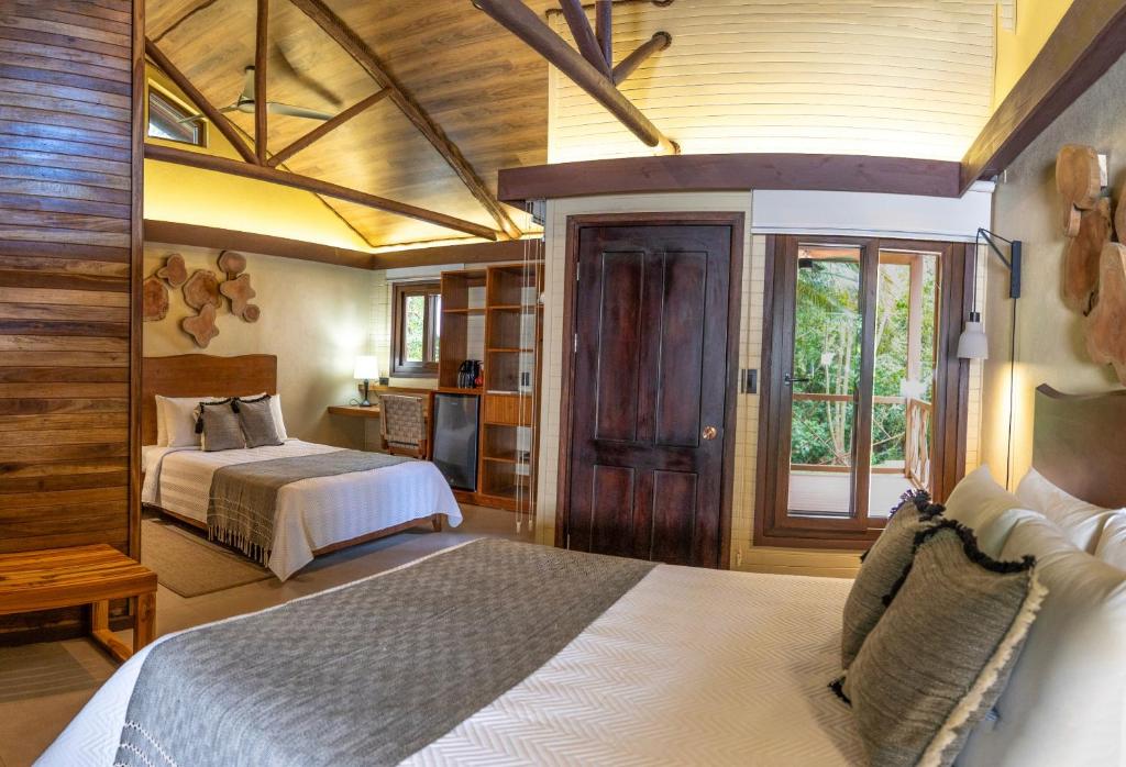 Spacious, rustic bedroom with two large beds, wooden accents, vaulted ceiling, a dark wooden door, bench, and large window revealing lush greenery outside. Natural light brightens the cozy, inviting space.