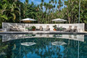 A tranquil pool reflects white umbrellas, lounge chairs, potted plants, and lush green trees under a bright blue sky. A white stone wall separates the pool area from the surrounding tropical foliage.