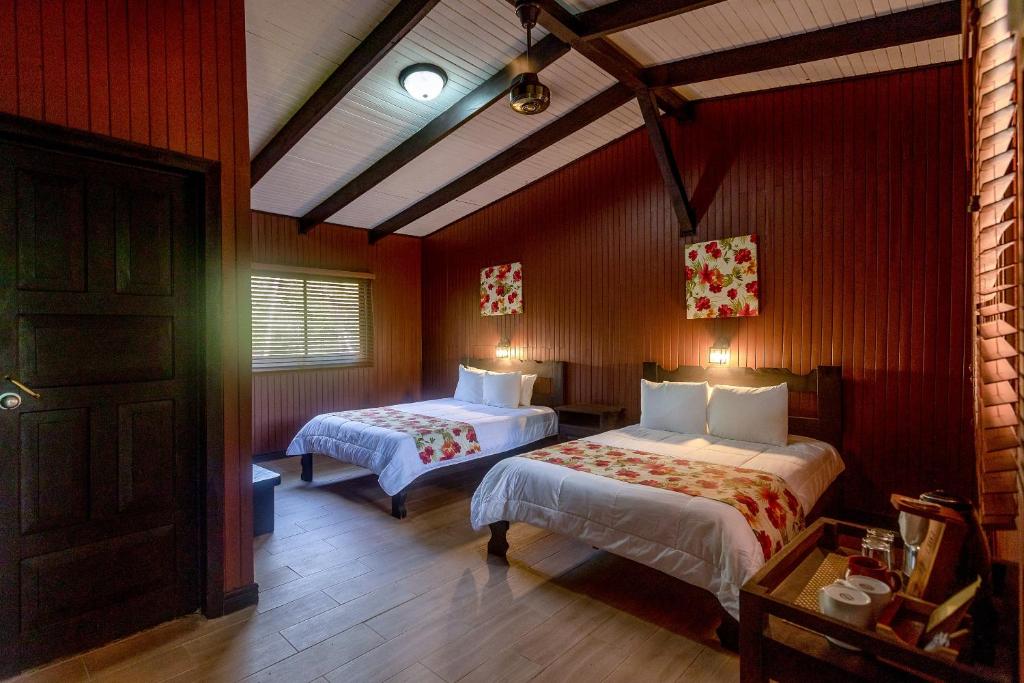 A cozy room with two double beds, white linens, and red floral accents. The walls are wooden, and there are two floral paintings above the beds. A window lets in natural light, and a tray with tea supplies is on a side table.
