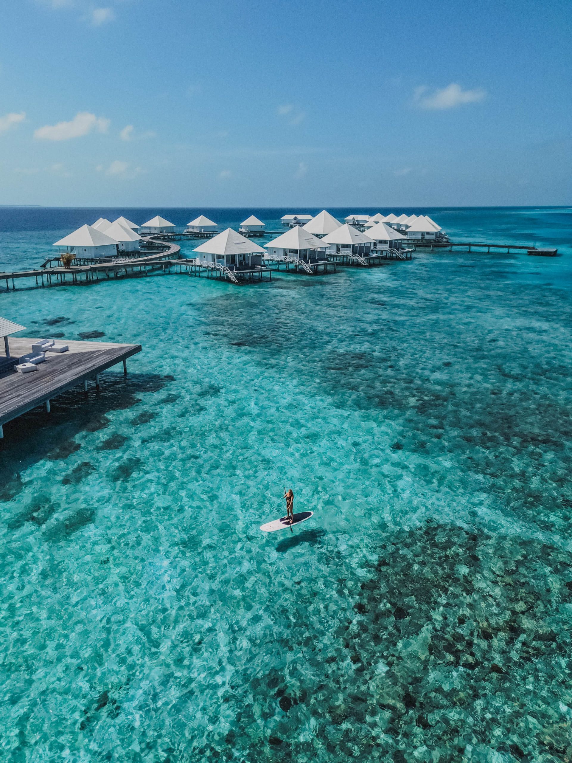A person stands on a paddleboard in clear turquoise water near overwater bungalows with white roofs, set against a bright blue sky and distant horizon.