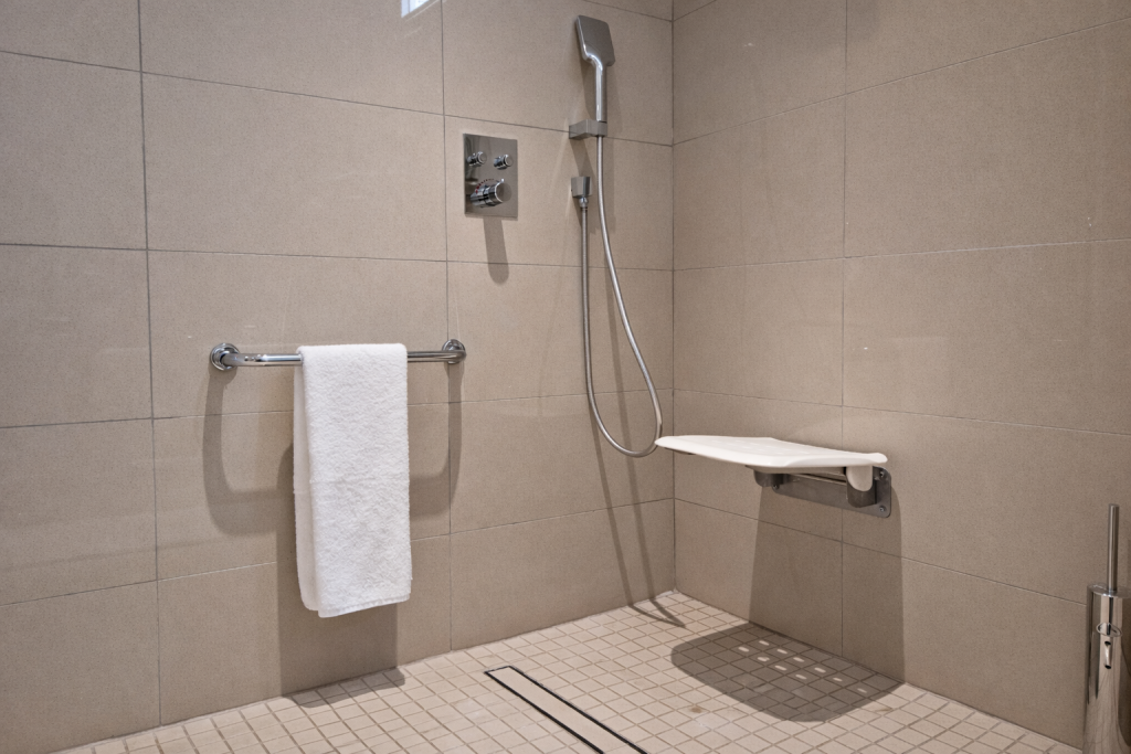 Accessible shower with level access entry, no raised threshold or lip. Floor finished in small non-slip mosaic tiles with linear drain. Wall-mounted fold-down shower
seat in white with chrome bracket on right wall. Handheld shower head on vertical sliding rail. Thermostatic shower controls with lever handles mounted at mid-height on rear wall. Horizontal chrome grab rail with towel on left wall. Partial vertical grab rail visible at far right edge. Wet room layout with large format tiles on walls.