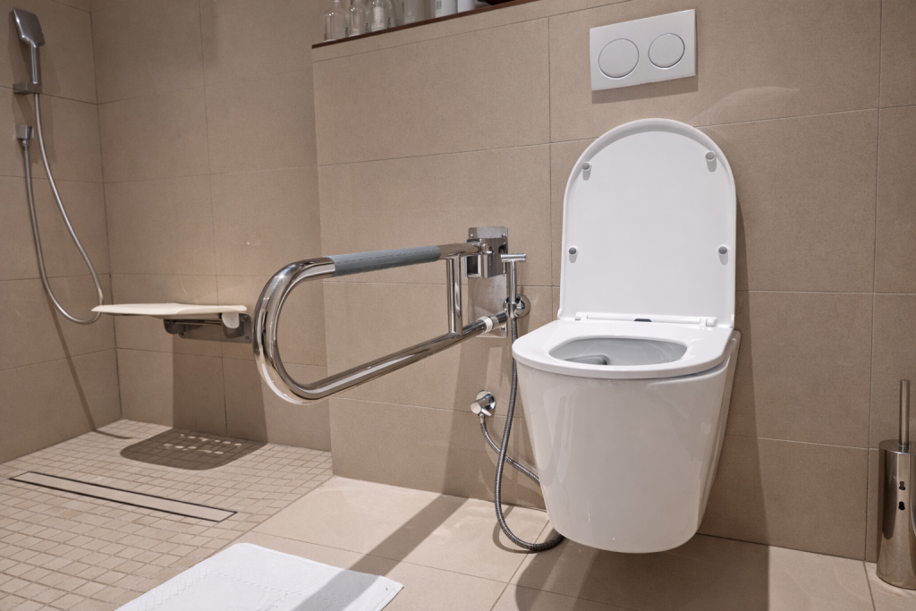 Wall-hung toilet close-up. Fold-down hinged grab rail mounted on left side of toilet (from seated position), chrome with grey foam grip. Rail extends full length
alongside toilet pan and folds up against wall when not in use. Wall-hung toilet with dual-button flush plate. Bidet spray attachment with flexible hose on left. Toilet seat open. No alarm cord visible in this image. Shower area with fold-down seat and non-slip floor visible
to left background.