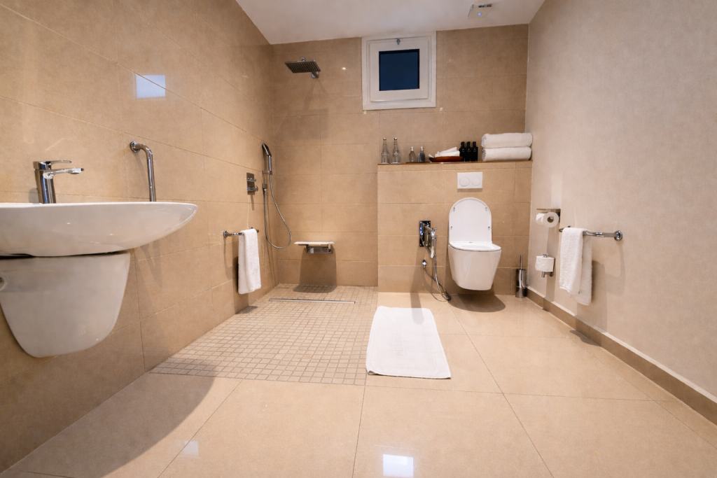 Accessible bathroom wide shot. Large open-plan layout with cream tiled walls and floor. Left: wall-hung basin with open under-sink clearance and lever tap — wheelchair approach possible. Centre: wet room shower zone with level access, fold-down seat, handheld shower head, overhead rain head, and non-slip mosaic tile floor with linear drain. Right: wall-hung toilet with cistern-less flush plate above and bidet attachment. No steps or raised thresholds anywhere in visible floor area. Generous clear turning space throughout. Small high-set window for ventilation.