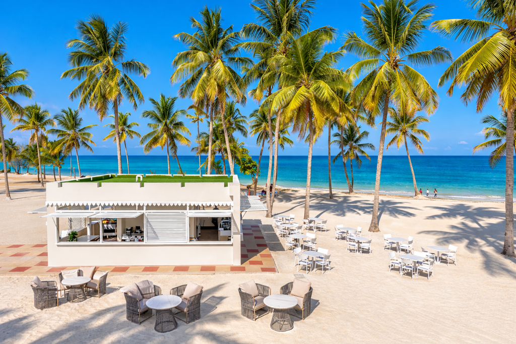 Aerial view of beach bar and terrace. White open-sided bar structure with scalloped canopy visible centre. Rattan lounge furniture on sand foreground. White dining tables and chairs on sandy terrace to right. Dense palm trees extending to white sand beach and turquoise ocean. Hotel building with arched architecture visible right background. Confirms large open beach with generous seating capacity.