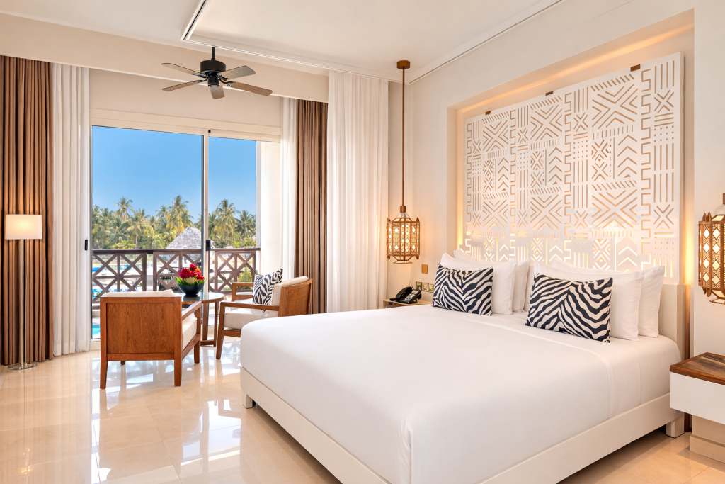 Hotel bedroom with large format cream tiled floor. King bed with white linen and geometric
patterned illuminated headboard panel. Pendant lantern bedside lights. Bedside telephone  and socket panel visible. Ceiling fan. Sliding glass doors opening to furnished balcony with two armchairs and small table, overlooking pool, palm trees and ocean. Floor lamp right. No steps visible between room and balcony.