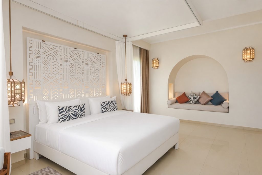 Hotel bedroom with large format cream tiled floor. King bed with white linen, geometric patterned headboard panel with backlit surround. Pendant lantern bedside lights. Built-in arched nook on right wall with cushioned seating in cream and coloured cushions. Sheer
curtain canopy framing. Balcony door with natural light beyond. Bedside table partially visible left.