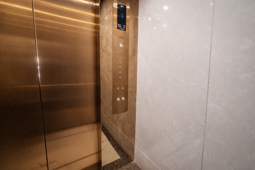 Lift interior control panel on marble-effect wall. Digital floor indicator display showing G (ground). Speaker/intercom grille below display. Approximately seven
raised circular buttons: one isolated at top, five floor selector buttons stacked vertically, two pairs at base (likely door open/close and alarm). Bronze lift doors to left. All buttons appear tactile and raised.