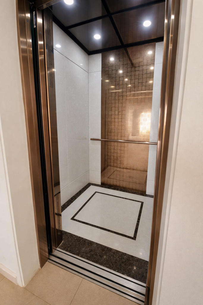 Lift interior with open doors. White panelled walls with bronze trim.
Handrail fixed horizontally to rear wall at mid-height. White marble floor with dark granite border inlay. Mirrored dark ceiling with recessed spotlights. Door opening appears wide. Space sufficient for wheelchair access.