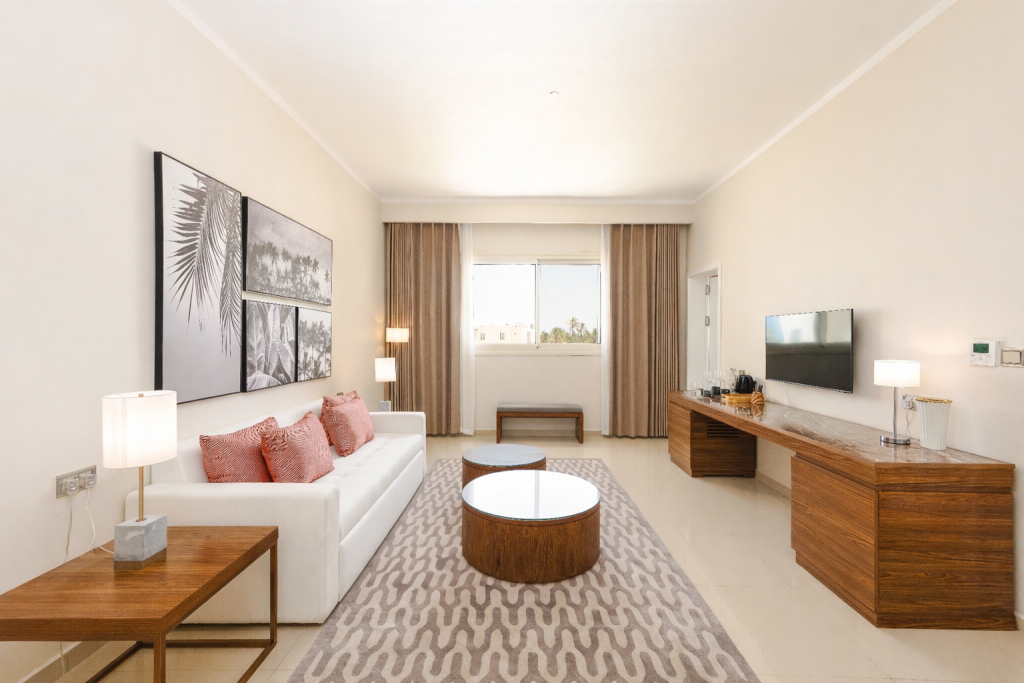 Suite sitting room with cream tiled floor and patterned area rug. White upholstered sofa with coral cushions. Circular timber coffee table. Dark timber TV and media unit with flat-screen,
tea tray, and kettle. Wall-mounted thermostat/climate control panel visible right. Tropical monochrome artwork triptych on left wall. Window with garden view and sheer curtains. Floor lamp. Separate room from bedroom.