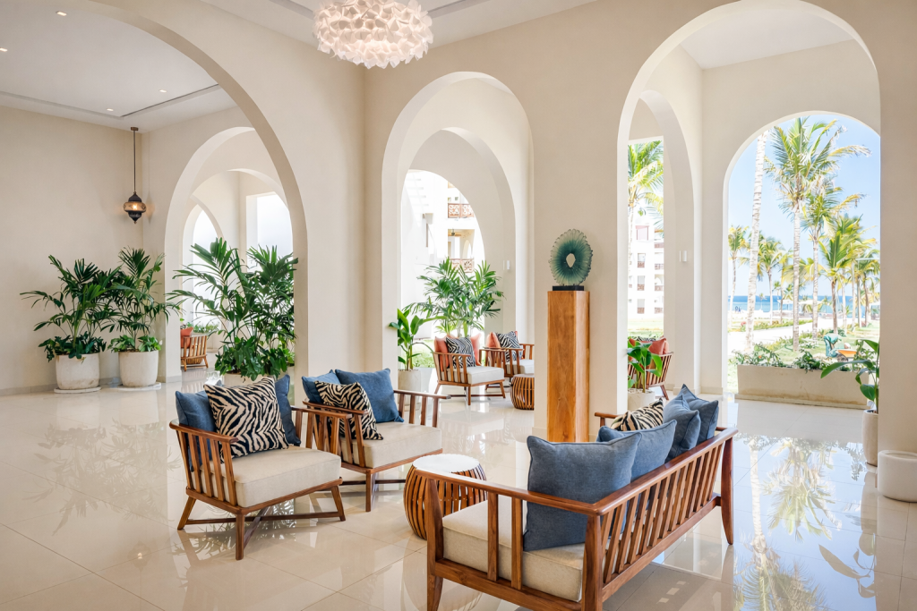 Open-sided hotel lobby lounge with series of tall arched openings. Polished white marble tile
floor. Timber-framed sofas and armchairs with zebra-print and blue cushions arranged in seating groups. Tropical potted plants throughout. Decorative sculpture on plinth. Direct views through arches to gardens, palm trees, and ocean. Pendant chandelier overhead.