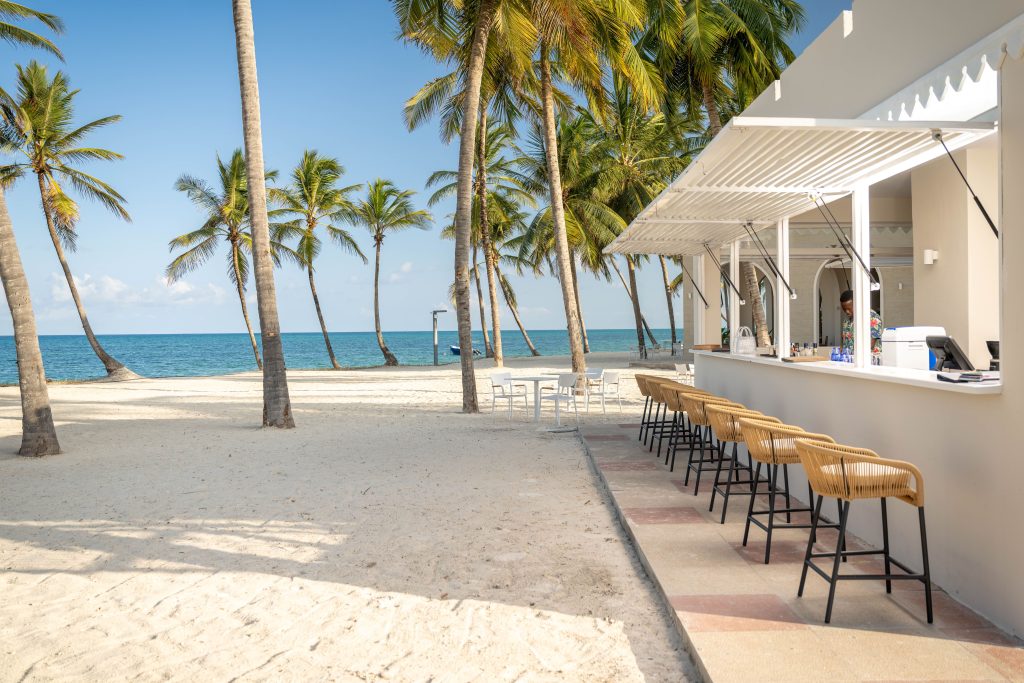 Beach bar with row of rattan-weave bar stools on high black metal frames along open service counter. Barman visible behind counter preparing drinks. Retractable louvred roof structure. White sand beach immediately adjacent with tables and chairs. Direct ocean views
and palm trees. Confirms bar seating on beach with table service area alongside.