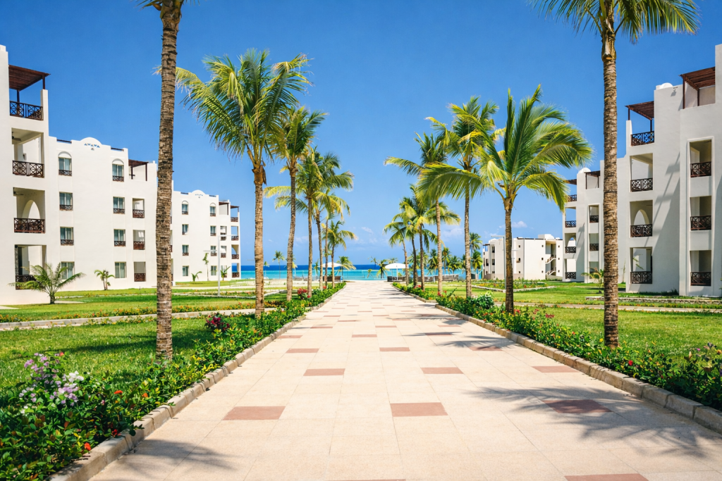 Wide paved central walkway between two white rendered accommodation blocks, three to
four storeys. Pathway lined with palm trees, manicured lawns, and flowering borders. Direct sightline to ocean and beach at far end. Arched window and balcony detailing on buildings. Flat paved surface throughout visible length — no steps on main approach route.