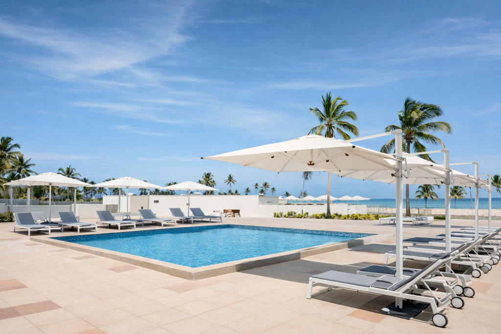 Rectangular outdoor swimming pool with blue mosaic tile finish. Pool terrace with white sun
loungers and large cantilever parasols on pale stone paving. Pool entry point visible at near edge — raised coping surround with no visible ladder or zero-entry steps from this angle. Palm trees and ocean visible beyond low boundary wall. Beach area with additional loungers in background.