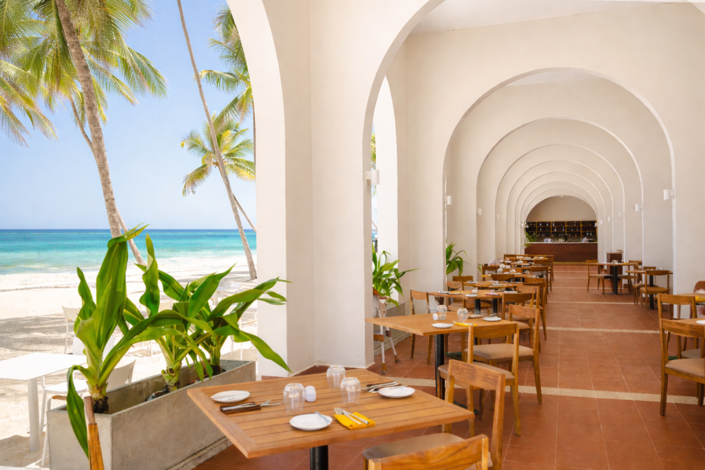 Open-sided beachfront restaurant with arched colonnade. Timber dining tables set for service with white plates and yellow napkins. Terracotta tile floor. Arches frame direct views of white sand beach, palm trees, and turquoise ocean. Bar/service area visible at far end of colonnade. Tropical planting at threshold between dining space and beach.
