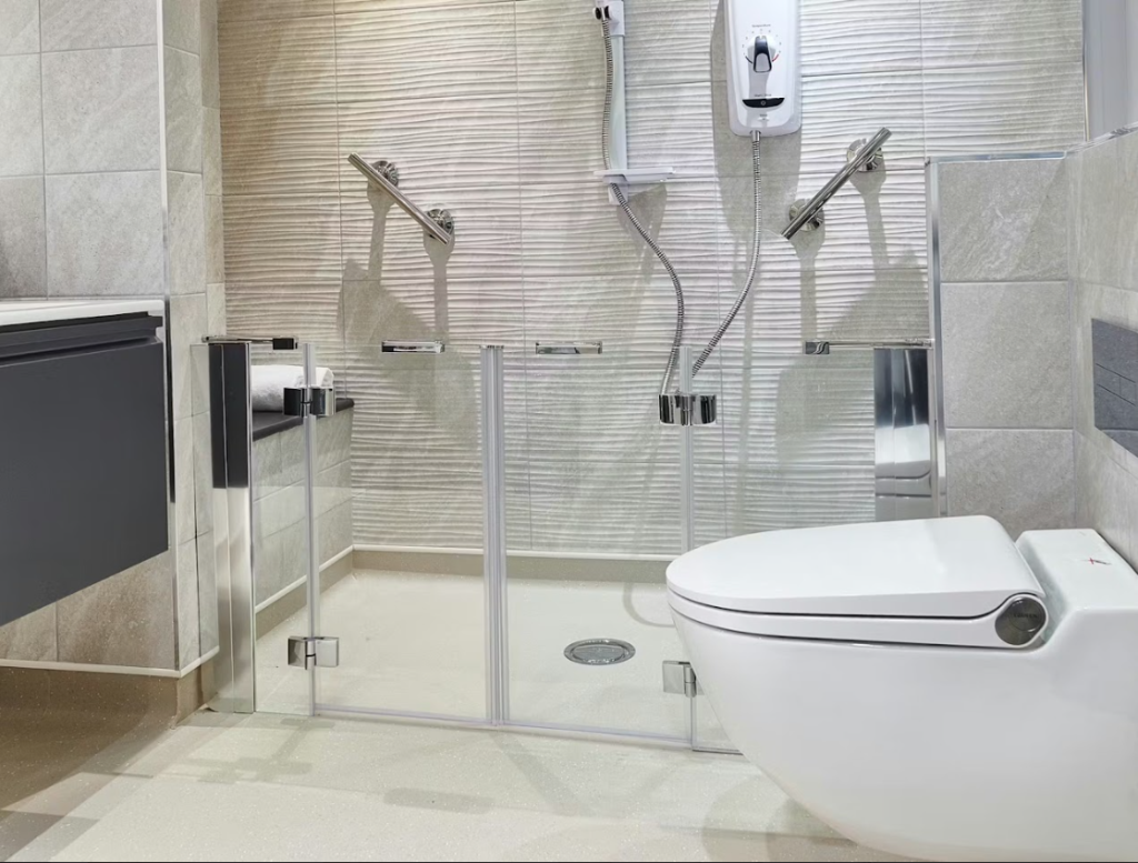 Glass-enclosed shower enclosure with bi-fold glass door panels and low threshold entry. Two angled chrome grab rails inside enclosure. Electric shower unit mounted high on rear wall with handheld shower head on flexible hose. Circular floor drain. Wall-hung smart toilet with integrated control panel to right of shower. Grey floating vanity unit to left. Textured wall tiles.