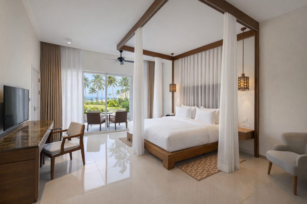 Hotel bedroom with large format cream tiled floor. Timber-framed four-poster king bed with
sheer white canopy drapes and upholstered headboard. Pendant lantern bedside lights. Flat-screen TV on dark timber media unit with desk and chair to left. Ceiling fan. Sliding glass doors opening to furnished balcony with ocean, palm trees, and garden view. Armchair in foreground right.