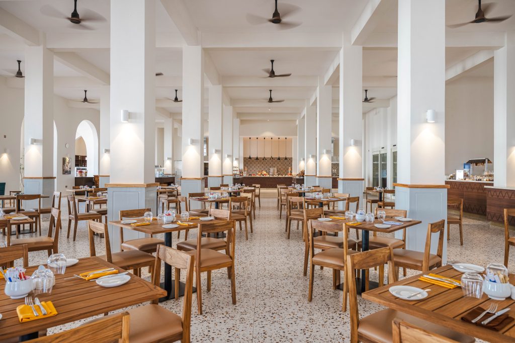 Spacious restaurant dining room with high white ceiling, multiple ceiling fans, and rows of structural white columns. Wooden tables set for dining with yellow napkins, glassware, and cutlery. Upholstered wooden chairs. Terrazzo-style floor. Buffet service area with pendant lighting visible at far end. Arched alcoves along left wall. Natural light from right.