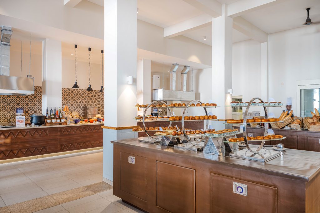 Large open-plan restaurant and buffet space with high white ceiling and ceiling fans. Long buffet counter with patterned tile splashback and pendant lighting in background. Island bread station in foreground with tiered displays of pastries, croissants, and rolls. Live cooking station visible right. Tiled floor with generous aisle space between counters.