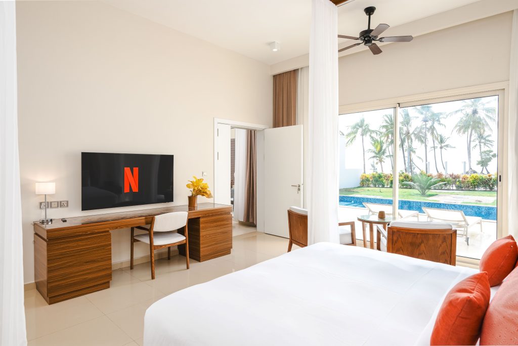 Hotel bedroom with king bed, cream tiled floor. Dark timber desk unit with flat-screen TV
displaying Netflix. Wooden desk chair. Ceiling fan. Floor-to-ceiling sliding glass doors opening to private terrace with pool, sun loungers, palm trees and ocean visible. Door to bathroom/corridor open on left wall.