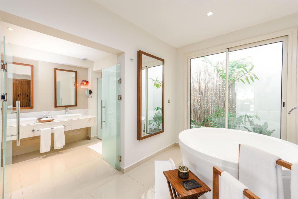 Large bathroom with freestanding oval bathtub positioned beside full-height glass doors opening to private garden. Glass-enclosed shower cubicle with chrome fittings centre-left. Double vanity with vessel sink, marble surround, and timber-framed mirrors. Cream tiled
floor throughout. Abundant natural light.