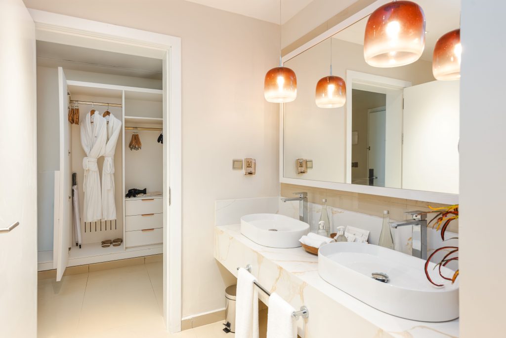 Open walk-in wardrobe showing hanging rail with robes and wooden hangers, lower drawer unit, and hair dryer. Smooth tiled floor. Adjacent bathroom area with double vessel sinks on marble countertop, large mirror above, amber pendant lights. Wall-mounted power sockets between wardrobe and vanity.