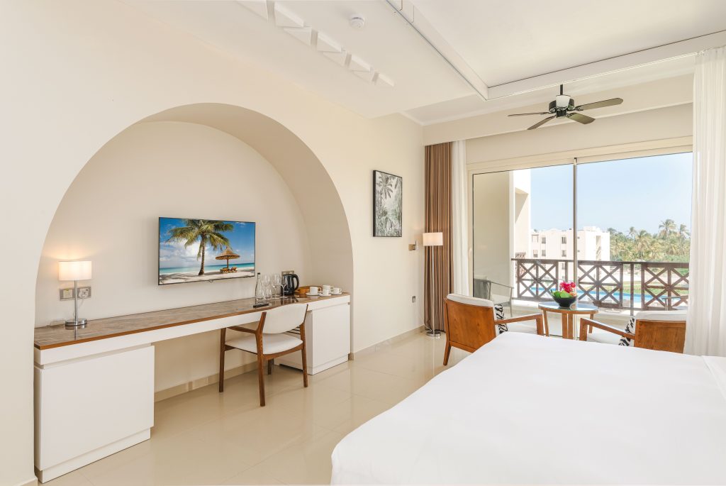 Hotel bedroom with smooth cream tiled floor and high ceiling. Arched alcove housing wall-mounted flat-screen TV above a built-in desk/counter with chair. Tea and coffee items visible on desk surface. Floor-to-ceiling sliding doors opening to furnished balcony with pool and garden view beyond. Ceiling fan. Bed partially visible foreground.