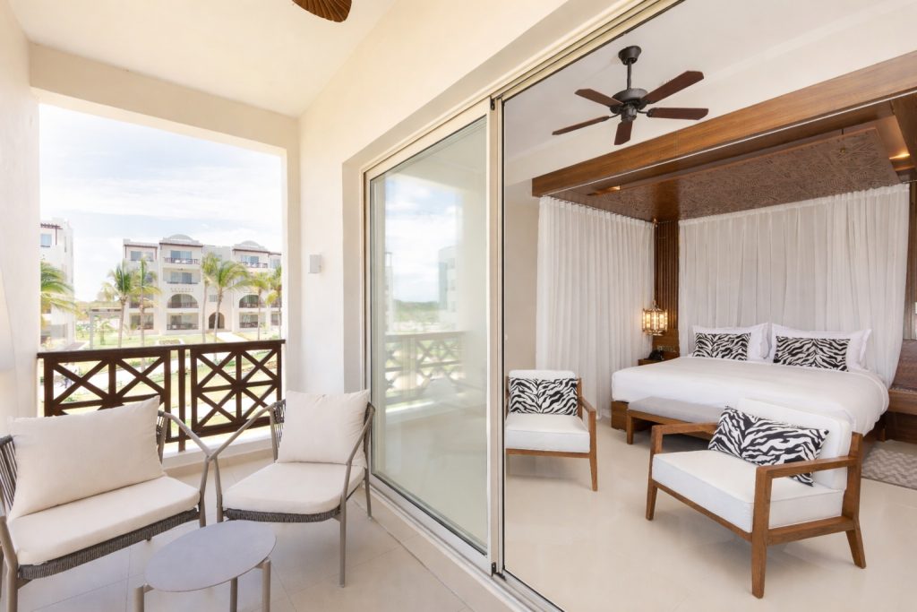 Private balcony with two white upholstered chairs and small round table. Full-height sliding glass doors opening to hotel bedroom beyond. Bedroom visible through glass showing timber-framed four-poster bed with sheer drapes, two armchairs, and ceiling fan. Dark timber cross-rail balcony balustrade. Resort buildings and palm trees visible in background. Balcony depth appears sufficient for comfortable seating.