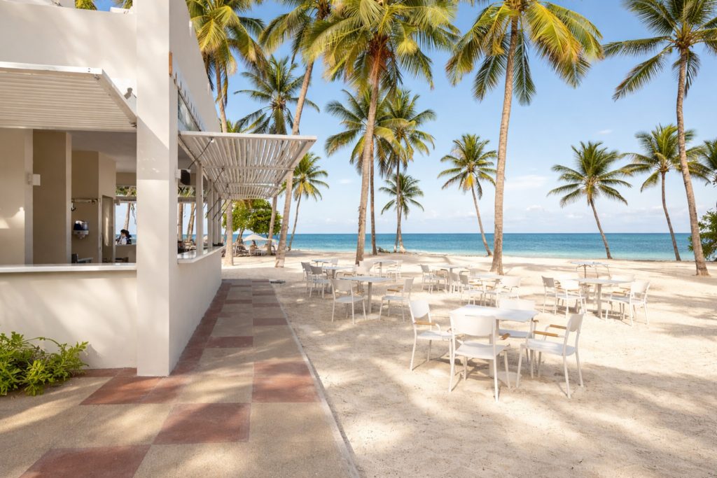 Open-sided beach bar with white rendered structure and louvred pergola roof. Service window with bar counter visible left. White plastic outdoor tables and chairs set out on paved and sandy terrace. Palm trees and turquoise ocean directly beyond. Confirms beach bar has sit-down dining as well as bar seating.