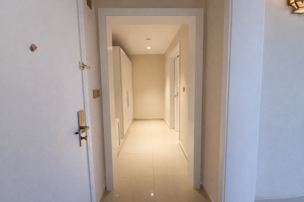 Hotel room entry viewed from corridor. Door open showing lever handle and electronic keycard reader mounted on door frame exterior. Door threshold appears flush with no raised lip. Internal entrance hallway with smooth cream tiled floor extends approximately 4–5 metres. Built-in wardrobe units on left wall. Recessed ceiling light. No steps or raised thresholds visible within view.
