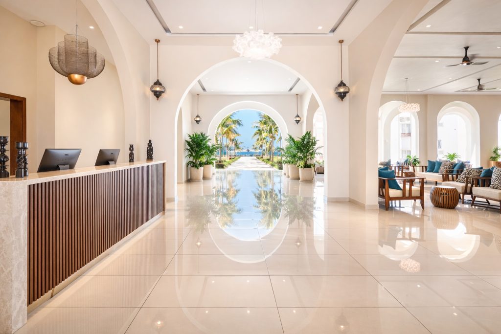 Hotel reception lobby with long timber-clad reception desk and two desktop screens on left. Polished cream tile floor with strong reflections. Central elliptical arch framing direct sightline through gardens and palm trees to ocean beyond. Arched side openings to right leading to lounge seating area with sofas and tub chairs. Decorative pendant chandeliers and Moorish-style hanging lanterns. Ceiling fan visible right.