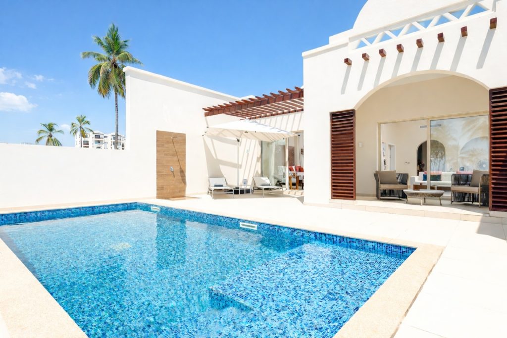 Private outdoor swimming pool belonging to a villa or suite. Rectangular pool with blue
mosaic tile. Pool terrace with sun loungers and retractable parasol. Outdoor shower on timber-clad wall. Sliding glass doors opening directly to interior living space. Covered outdoor seating area to right with rattan furniture. White rendered villa architecture with
timber pergola and louvre shutters.
