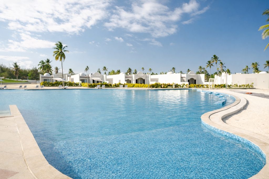 Large freeform outdoor swimming pool with blue mosaic tile finish. Curved stepped entry into pool visible right — three or four shallow steps descending into water with no handrail visible. Pool surrounds with sun loungers and parasols. White resort buildings and palm trees in background. Open sky.