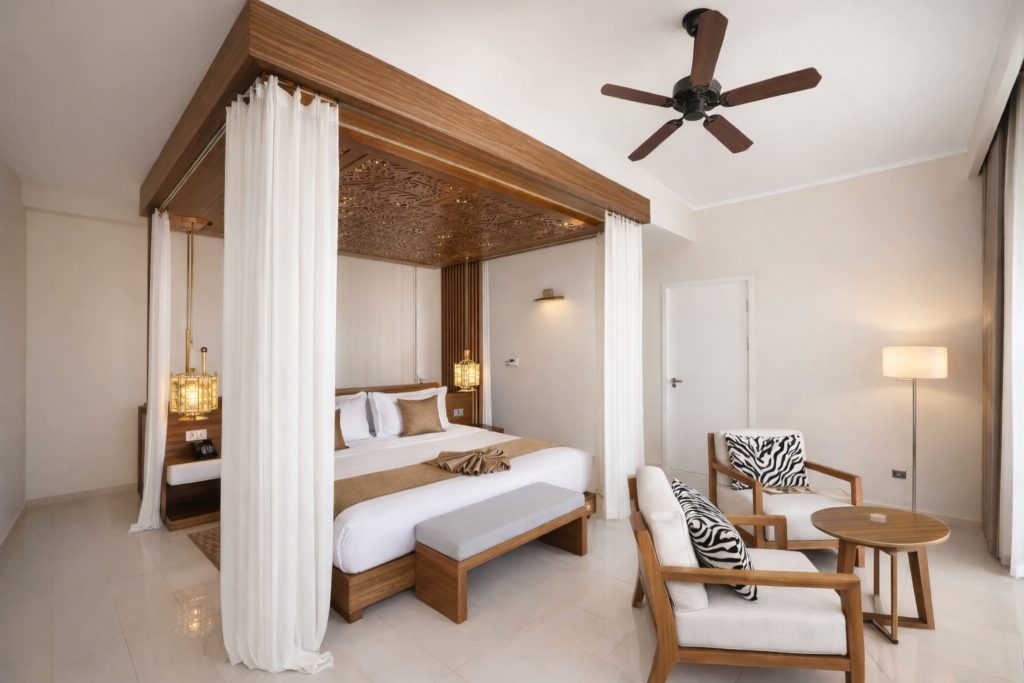 Hotel bedroom with large-format cream tiled floor. Solid timber-framed four-poster king bed
with carved ornamental canopy ceiling insert and sheer white drapes. Pendant lantern bedside lights. Timber integrated headboard unit with bedside tables. Upholstered bench at foot of bed. Two wooden armchairs with zebra-print cushions and round side table. Ceiling fan. Door to corridor visible right.