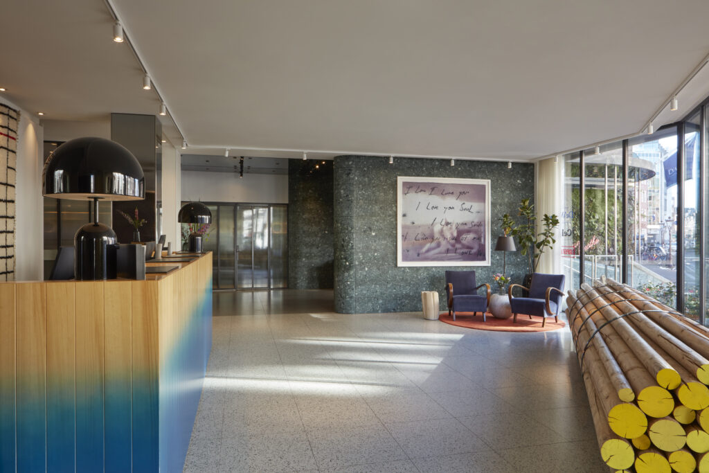 Kimpton de Witt reception area with light wood desk and oversized black table lamps, seating with blue velvet armchairs on terrazzo flooring beside the green stone column.