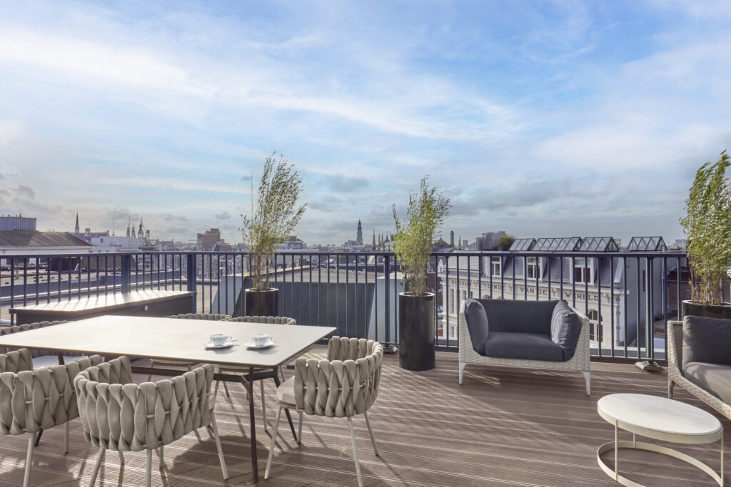 Penthouse de Witt private rooftop terrace with timber decking and metal railings, dining setting with woven chairs and lounge area with armchairs, view across Amsterdam rooftops to historic spires.