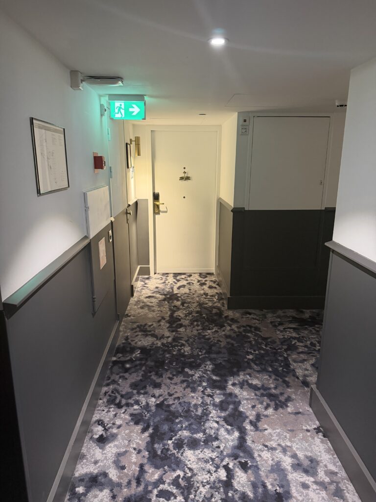 Corridor outside accessible bedroom showing room door with illuminated exit sign and patterned carpet.