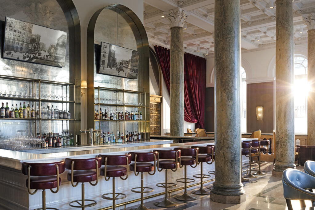Cafe Riggs bar at Riggs Washington DC. White marble counter with burgundy leather bar stools. Brass and glass shelving behind the bar holds spirits and stemware. Marble columns separate the bar from the seating area. Black and white framed historical photographs of Washington DC mounted above the back bar. Natural daylight from arched windows on the right.