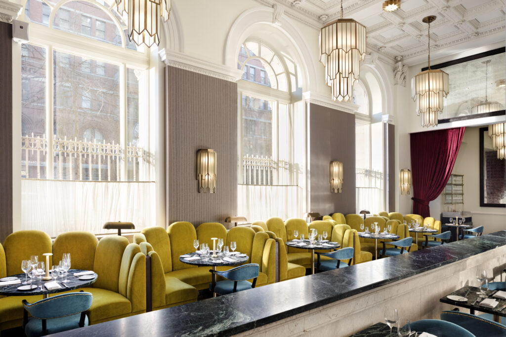 Main dining room at Cafe Riggs, the all-day restaurant on the ground floor of Riggs Washington DC. Mustard yellow velvet banquettes line the marble bar counter. Round marble tables paired with blue velvet chairs. Tall arched windows on the left wall above wood panelling. Art deco brass and glass chandeliers hang from a moulded white ceiling.